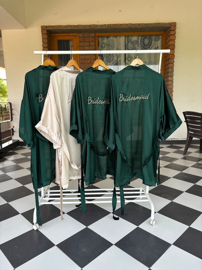 May include: Four satin robes hanging on a rack. Three robes are dark green and one is cream. The green robes have the word "Bride" or "Bridesmaid" embroidered on the back in gold thread.