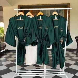 May include: Four dark green satin robes with gold embroidery hanging on a white rack. The robes have a tie belt and are embroidered with the word "Mrs." on the back.