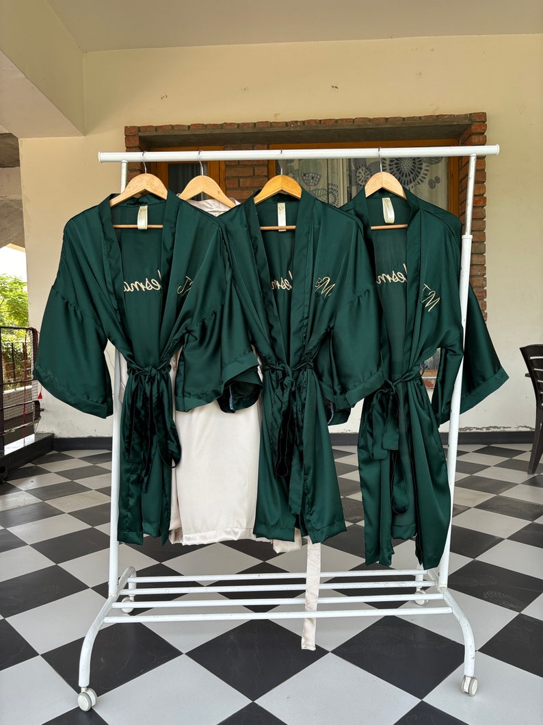 May include: Four dark green satin robes with gold embroidery hanging on a white rack. The robes have a tie belt and are embroidered with the word "Mrs." on the back.