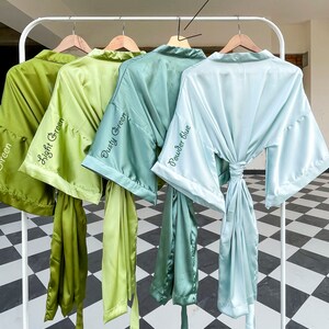K&ouml;nnte beinhalten: Vier Satin-Badem&auml;ntel in Gr&uuml;n- und Blaut&ouml;nen h&auml;ngen an einem wei&szlig;en Gestell. Die Badem&auml;ntel sind mit "Light Green", "Dusty Green" und "Powder Blue" beschriftet. Jeder Bademantel hat einen passenden Bindeg&uuml;rtel.