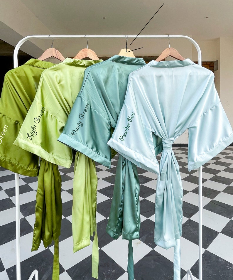 K&ouml;nnte beinhalten: Vier Satin-Badem&auml;ntel in Gr&uuml;n- und Blaut&ouml;nen h&auml;ngen an einem wei&szlig;en Gestell. Die Badem&auml;ntel sind mit "Light Green", "Dusty Green" und "Powder Blue" beschriftet. Jeder Bademantel hat einen passenden Bindeg&uuml;rtel.