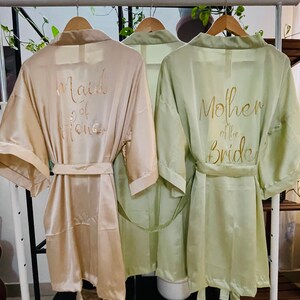 May include: Two satin robes hanging on a rack. The robe on the left is beige with the text "Maid of Honor" in gold lettering. The robe on the right is sage green with the text "Mother of the Bride" in gold lettering.