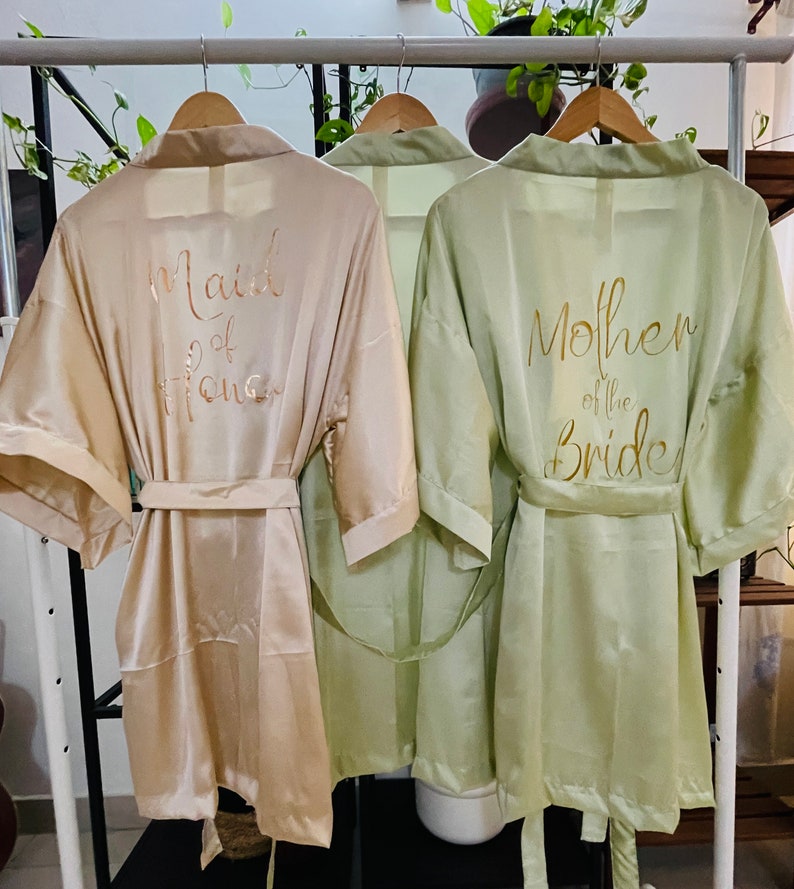 May include: Two satin robes hanging on a rack. The robe on the left is beige with the text "Maid of Honor" in gold lettering. The robe on the right is sage green with the text "Mother of the Bride" in gold lettering.