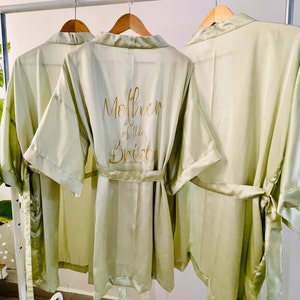 May include: Three sage green satin robes with gold lettering. The middle robe has the text "Mother of the Bride" on the back.
