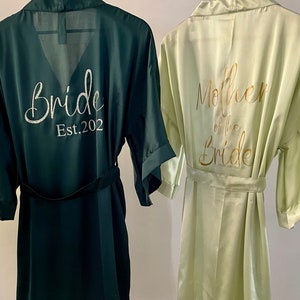 May include: Two satin robes, one dark green with "Bride Est. 2022" in gold lettering, and the other light green with "Mother of the Bride" in gold lettering.