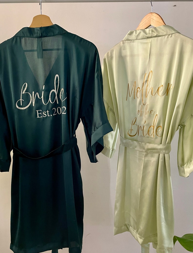 May include: Two satin robes, one dark green with "Bride Est. 2022" in gold lettering, and the other light green with "Mother of the Bride" in gold lettering.