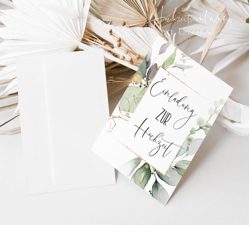 May include: A white card with gold lettering that says "Einladung zur Hochzeit" with a green watercolor floral design. The card is on a white surface with a white envelope.