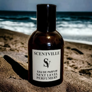 May include: A brown glass bottle of perfume with a white label that says "SCENTVILLE" and "S V" in a stylized font. The label also says "EAU DE PARFUM NEXT LEVEL PERFUMERY". The bottle is sitting on a sandy beach.