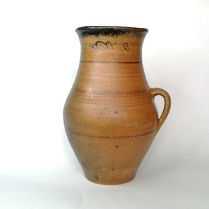 Może przedstawiać: Rustykalny, brązowy ceramiczny dzban z uchwytem. Dzban ma szeroką podstawę, która zwęża się ku węższej szyi i rozszerzonej krawędzi. Ciemnobrązowe paski otaczają korpus. Obrzeże jest ciemnobrązowe.