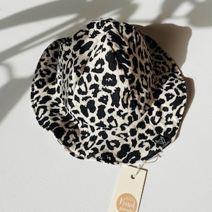 Könnte beinhalten: Eimerhut mit Leopardenmuster in Schwarz und Creme. Der Hut hat eine breite Krempe und ein Etikett mit der Aufschrift "100% Hand gemacht". Der Hut ist aus einem weichen Stoff gefertigt und eignet sich zum Sonnenschutz.