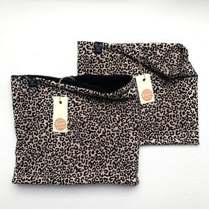 Könnte beinhalten: Zwei braune und schwarze Leopardenmuster-Halswärmer mit schwarzem Fleece-Futter. Ein Halswärmer ist gefaltet und der andere liegt flach. Beide Halswärmer haben ein kleines braunes Etikett mit dem Text "100% Handgefertigt" darauf.