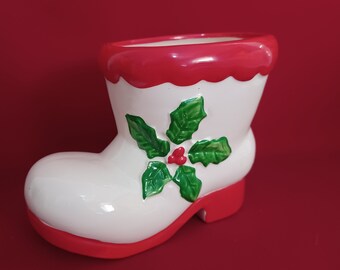 May include: A ceramic Christmas boot planter. The boot is white with a red scalloped top and red sole. Green holly leaves and red berries decorate the side. The planter is set against a red background.