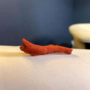 K&ouml;nnte beinhalten: Ein kleines, strukturiertes, rot-oranges Holzst&uuml;ck, m&ouml;glicherweise ein dekoratives Element oder ein Naturgegenstand. Das Holz hat eine raue Oberfl&auml;che und eine unregelm&auml;&szlig;ige Form, die auf einer wei&szlig;en Oberfl&auml;che mit dunklem blauem Hintergrund liegt.