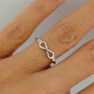 May include: A silver-coloured ring featuring an infinity symbol design centred on the band. The infinity loop is accented with two small, clear, round-cut stones, one positioned at each end of the symbol, set into the polished metal band.