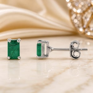 May include: A pair of stud earrings featuring rectangular emerald-cut green gemstones set in silver-toned metal. One earring is shown from the front, while the other is displayed from the side, revealing the post and the circular butterfly back closure.