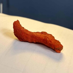 K&ouml;nnte beinhalten: Ein gebogenes, rot-oranges Holzobjekt mit strukturierter Oberfl&auml;che. Das Objekt hat eine nat&uuml;rliche, organische Form und scheint ein St&uuml;ck Holz oder ein &auml;hnliches Material zu sein. Die Farbe ist durchgehend, ohne sichtbare Variationen oder Muster.