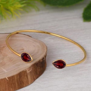 Könnte beinhalten: Ein goldfarbenes, offenes Armband mit zwei tropfenförmigen, tiefroten Granat-Edelsteinen an den Enden. Das Metallband ist dünn und kreisförmig gestaltet, sodass es um das Handgelenk getragen werden kann, wobei sich die beiden Steine gegenüberliegen.