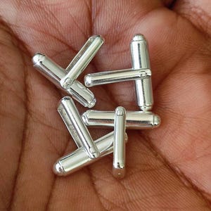 May include: A close-up of several shiny silver cufflinks. The cufflinks have a classic design with a rounded barrel shape and a connecting bar. The metal reflects light, creating a bright and polished appearance. The cufflinks are displayed on a skin-toned background.