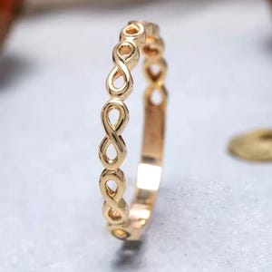 Puede incluir: Un anillo de tono dorado que presenta un diseño de banda continua compuesto por símbolos de infinito repetidos. El metal tiene una superficie pulida y reflectante, y la estructura del anillo está formada por los bucles interconectados.