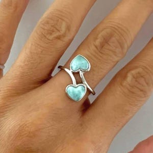 May include: A silver ring featuring two heart-shaped light blue stones with a marbled texture. The band has a bypass design where the two hearts are positioned at different heights, creating an open, asymmetrical look on the finger.