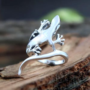 May include: A polished silver-toned ring shaped like a lizard. The lizard's body forms the band of the ring, with its head and feet extending upwards. The metal surface is smooth and reflective, showcasing the detailed form of the reptile.