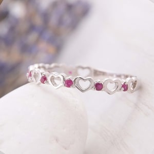 Könnte beinhalten: Ein silberfarbener Ring mit einem sich wiederholenden Muster aus offenen Herzformen, die sich mit kleinen, runden, leuchtend pinkfarbenen Steinen abwechseln. Das Band hat ein filigranes, durchgehendes Design mit polierter Metalloberfläche.
