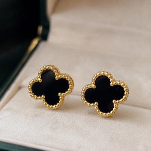 May include: A pair of stud earrings featuring a four-leaf clover shape with a smooth black center inlay. The clover motif is outlined by a gold-toned beaded border, creating a textured edge around the dark, solid-colored central element.