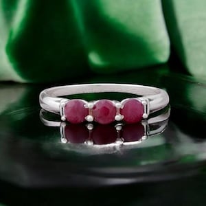 Könnte beinhalten: Ein silberfarbener Ring mit drei runden, tiefroten Edelsteinen, die horizontal in einer Reihe angeordnet sind. Die Ringschiene ist glatt und poliert und zeigt ein schlichtes Design, bei dem die drei Steine markant auf der Oberseite sitzen.