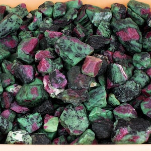 Pode incluir: Uma coleção de pedras brutas e não polidas de rubi em zoisite, apresentando uma mistura de cristais de rubi magenta vibrantes incrustados numa matriz mineral de zoisite verde e preta manchada, criando uma superfície densa e texturizada.