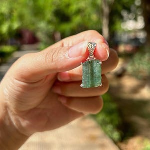 May include: A raw green tourmaline crystal pendant set in a silver-toned metal cap with a textured bail. The gemstone features a vertical, columnar structure with natural striations and a translucent, light green hue, suspended from a metal loop.