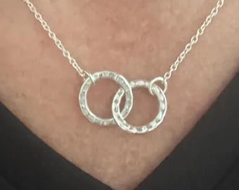 May include: A silver-toned necklace featuring two interlocking circular pendants with a hammered, textured finish. The delicate chain connects to the sides of the rings, creating a linked design that rests against the collarbone area.