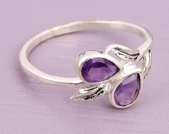 May include: A silver ring featuring two pear-shaped purple amethyst gemstones set in a metal band. The setting includes decorative silver leaf-like accents that frame the two stones, creating a nature-inspired design on the band's front section.