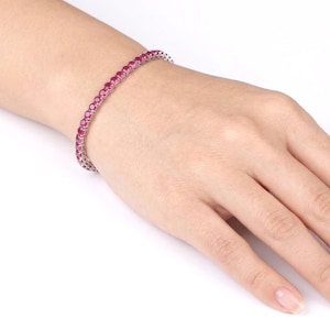 Könnte beinhalten: Ein Tennisarmband mit einer durchgehenden Reihe runder, leuchtend pinker Edelsteine, die in ein poliertes, silberfarbenes Metallband gefasst sind. Das Armband umschließt das Handgelenk und zeigt ein einheitliches Design mit sicher gefassten Steinen.