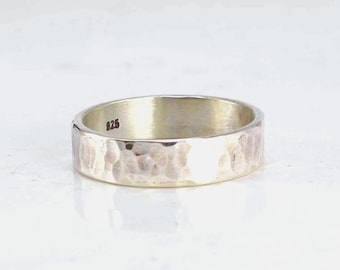 May include: A silver-colored band ring with a hammered, textured exterior finish. The interior of the ring features a clear stamp reading 925, indicating the metal composition. The ring has a flat profile and a polished, reflective metallic surface.