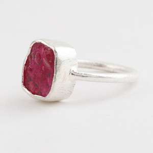 May include: A silver ring featuring a raw, uncut, deep pinkish-red gemstone set in a textured metal bezel. The band is smooth and rounded, showcasing a metallic finish that contrasts with the irregular, organic shape of the vibrant stone.
