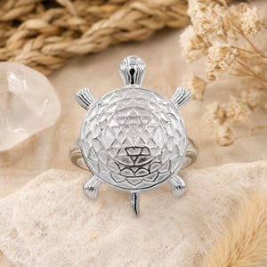 Op de afbeelding: Een zilverkleurige ring met een schildpadvormig middendeel. Het schild van de schildpad is gegraveerd met een geometrisch sterpatroon, terwijl de kop, vier vinnen en staart uitsteken vanaf het ronde lichaam. Het metaal heeft een glanzende afwerking.