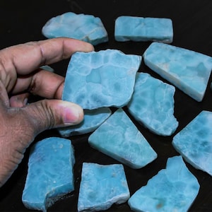 May include: A collection of flat, polished Larimar stone slabs featuring a distinct sky-blue color with white, cloud-like marbling patterns. The stones are irregular in shape, ranging from rectangular to trapezoidal, with smooth surfaces.