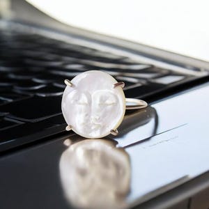 May include: A ring featuring a circular, iridescent white mother-of-pearl inlay carved with a serene, stylized human face. The round shell piece is secured by four small metal prongs onto a thin, polished metallic band that encircles the finger.