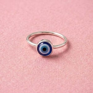 Puede incluir: Un anillo de tono plateado con un amuleto circular de ojo turco como pieza central. El diseño muestra anillos concéntricos de color azul oscuro, azul claro y blanco, con una pupila negra sólida en el centro, sobre una banda metálica pulida.