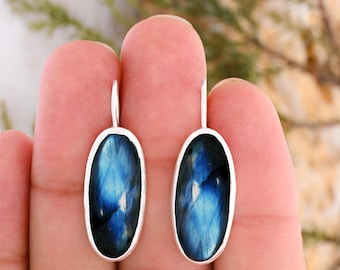 May include: A pair of oval-shaped earrings featuring polished labradorite gemstones with iridescent blue flashes. Each stone is set in a smooth silver-toned metal bezel with a simple hook attachment at the top, showcasing a natural stone texture.
