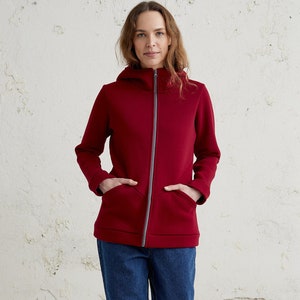 Könnte beinhalten: Eine rote Kapuzenjacke mit Reißverschluss und zwei Eingrifftaschen. Die Jacke wird über einer blauen Jeans und weißen Turnschuhen getragen.