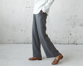 May include: A woman wearing a white linen top and grey wide-leg trousers. She is standing in front of a white textured wall and is looking at the camera.