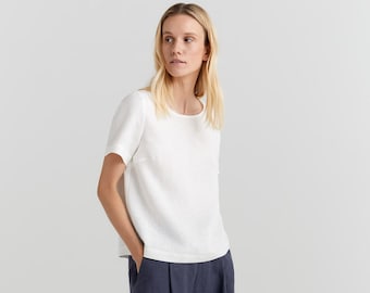 May include: A white linen top with short sleeves and a relaxed fit paired with dark blue linen trousers. The model is wearing grey sandals.