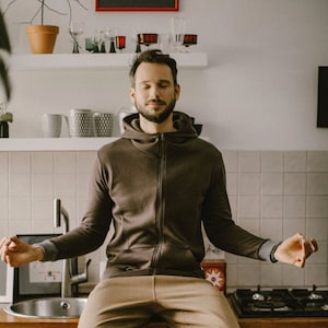 Może przedstawiać: Mężczyzna siedzi na blacie kuchennym z zamkniętymi oczami, medytując. Ma na sobie brązową bluzę z kapturem i beżowe spodnie. Kuchnia ma białe płytki na ścianie i stalowy zlew.