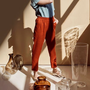 May include: A woman wearing a light blue shirt and rust-colored pants stands in a room with sunlight streaming through the windows. She is surrounded by clear glass vases of various shapes and sizes.