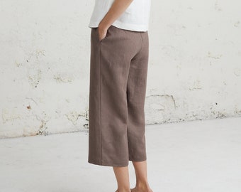 May include: A woman wearing a white sleeveless top and brown linen trousers. She is standing in front of a white wall.