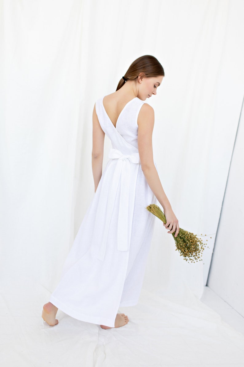 May include: A white linen dress with a V-neckline and a tie at the waist. The dress is flowing and has a relaxed fit. The model is holding a bouquet of dried flowers.