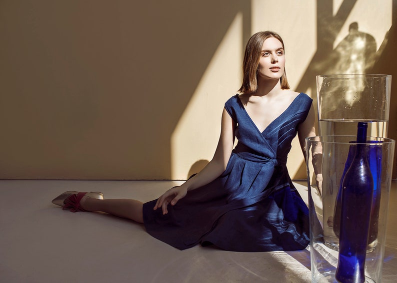 May include: A woman in a blue dress sits on the floor with her legs outstretched. She is looking up and to the right. There are two clear glass cylinders behind her, one containing a blue glass bottle.