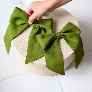 Può includere: Due fiocchi di lino verde sono appoggiati su una scatola rotonda beige. I fiocchi sono di una tonalità verde vibrante, con code lunghe e fluenti. La scatola ha una superficie strutturata. I fiocchi sono probabilmente per confezioni regalo o scopi decorativi.