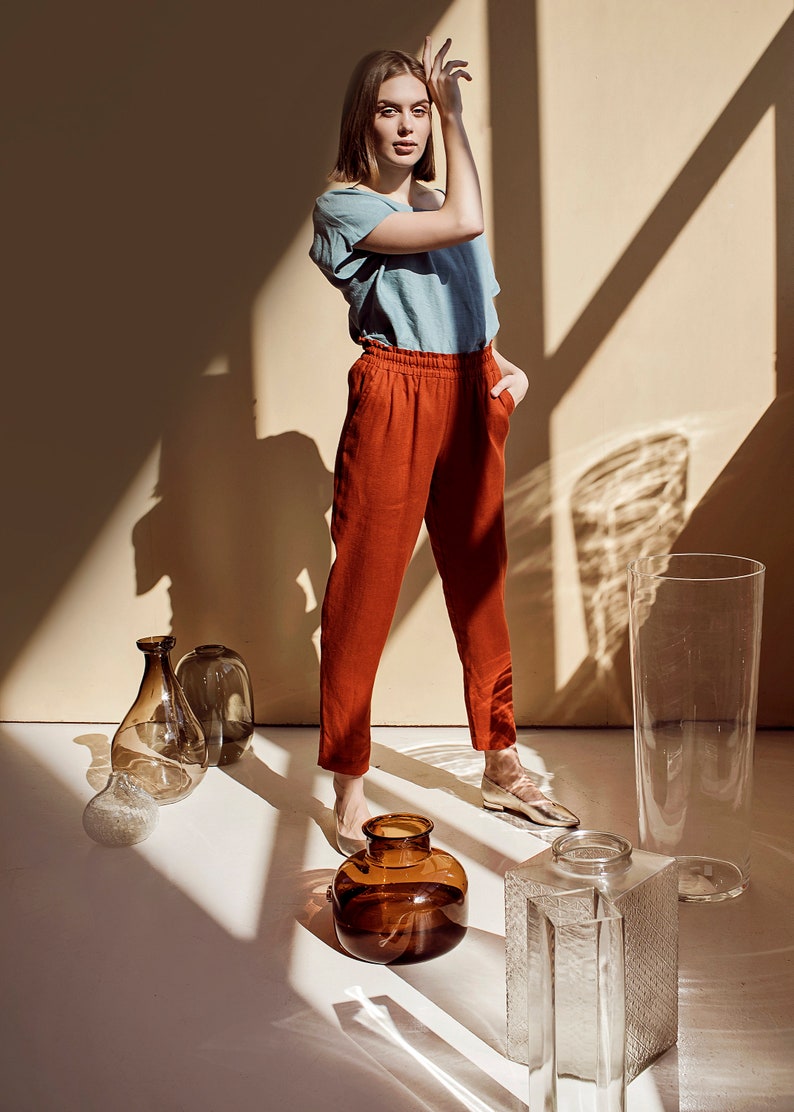 May include: A woman wearing a light blue shirt and rust-colored pants stands in a room with sunlight streaming through the windows. She is surrounded by clear glass vases of various shapes and sizes.